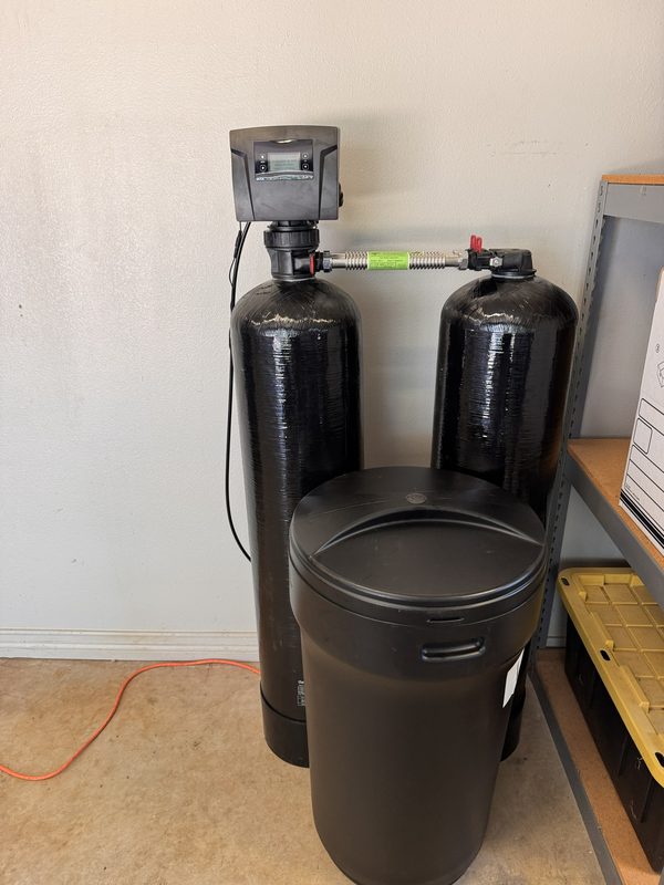 Whole-home water treatment system with two tall black media tanks, a water softener and a carbon filter, next to a black brine tank