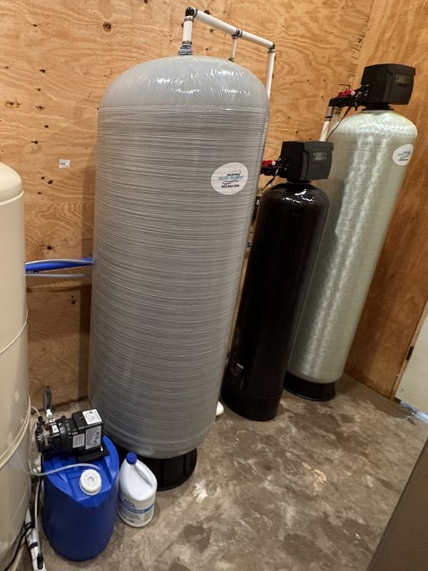 Indoor well water treatment system in a utility room: a chemical feed pump on a solution drum, a tall retention tank, and two filter tanks